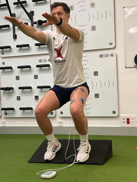 Basas Spanish Squat to strengthen patellar tendon:Person squats arms ahead with electrical stimulation electrodes on L thigh